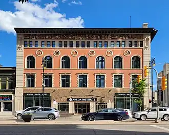 The front of a four storey building. On the main floor, the facade is brown tile. Floors above that it is terracotta-colored stucco. Just below the roofline is a detailed mosaic of many characaters represented in flat, egyptian-style design. Just below the fourth floor windows are decorative circles containing more mosaics. The over all design is very busy and detailed, while being symmetrical and solid.