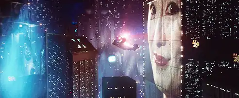 A still frame from the film Blade Runner. A flying car flies between skyscrapers in a futuristic looking city. The buildings are covered with lights and advertising video billboards.