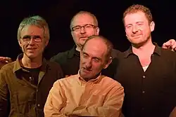 Four men, three behind and one at the front in a darkened jazz club , looking at the camera.