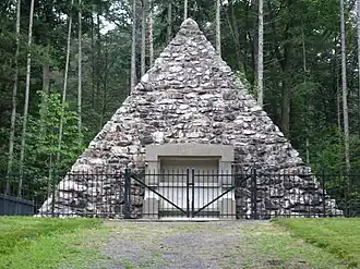 A fieldstone pyramid surrounded by a fence with pine trees in the background