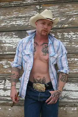 Portrait photo of transgender porn actor Buck Angel, standing in front of a wood panel building, smouldering at the camera, wearing a cowboy hat, an open button-down shirt, blue jeans and a large gold belt buckle. He has tattoos on his arms, chest, and stomach. He holds a cigar in one hand, and has his other thumb in the pocket of his jeans.