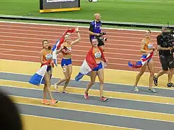 Photo of four athletes carrying Dutch flags, walking alongside a photographer and a cameraman, next to a red athletics track
