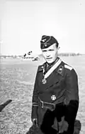 Black-and-white portrait of a standing man wearing a cap and black military uniform with an Iron Cross displayed at his neck.