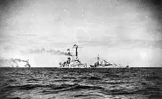 Two large warships steaming in opposite directions close together, with several other vessels are in the distance.