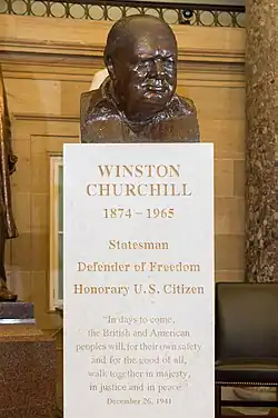 A brown bronze bust of Sir Winston Churchill. e has a smug look on his face, and is looking forward. On the white marble pedestal are the words "WINSTON CHURCHILL, 1874-1965, STATESMAN, DEFENDER OF FREEDOM, HONORARY U.S. CITIZEN".