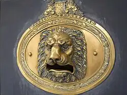 A bronze lion's head upon an oval shield beneath the Arms of Spain; the lion's mouth is a wide opening for letters.