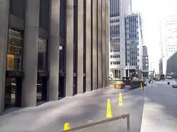 The recessed plaza surrounding the CBS Building, looking west along 53rd Street. The plaza has gray pavement and is several steps below the street. There are yellow cones in the plaza. To the left is the CBS Building, and to the right is the sidewalk on 53rd Street.
