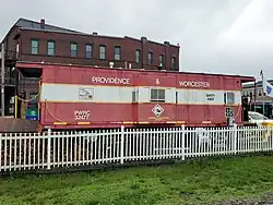A railroad caboose parked on a siding behind a white picket fence. It reads "PROVIDENCE & WORCESTER" on its side.