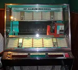 Blue and white Jukebox. Sign at the top of the machine reads "EP ALBUM RECORDS" with "SHOW TUNES JAZZ MOOD MUSIC" written to the left and "OLD FAVORITES CLASSICS DANCE MUSIC" written to the right. "Select-O-Matic Two-Hundred" is printed on the glass. A line of horizontal 45 records can be seen, as well as a rectangular Seeburg logo on the right side of the machine. Above and below the line of 45s a white catalogue can be seen. Between columns of the catalogue are four blue, black, and white illustrations. Clockwise starting with the top left illustration these depict: two men in suits playing horns, a woman sitting on a couch clutching her knees, a man in a vest dancing with a woman in a long dress, and a singer at a microphone with two floating 45s next to her head, accompanied by two men playing horns. Placards below the bottom row of the catalogue are visible, reading "HIT TUNES", "Rhythm and Blues", and "Country and Western". Buttons for selecting the 45s below the catalogue from left to right are lettered A through K, then numbered 1 through 9, then 0, then lettered L through V. The lettered buttons are red, and the numbered letters are green. A coin slot is on the right side of the buttons, reading "COINS NICKELS DIMES QUARTERS UP TO ONE DOLLAR TOTAL OR A HALF DOLLAR".