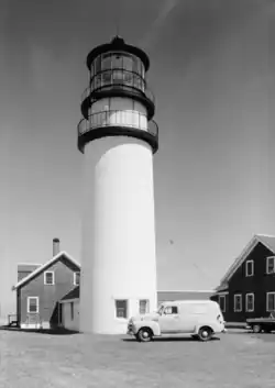 High-resolution black and white photo of the lighthouse and keeper's house. Parked in front of the lighthouse is a 1950s van, with a military emblem and the words "United States Coast Guard" stenciled on the driver's door, and on the side of the van is a military advertisement that reads "The U.S. Coast Guard NEEDS MEN! Join Now! No waiting".