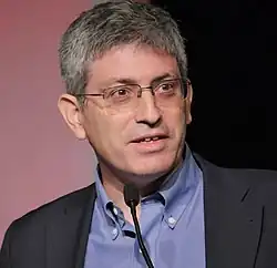 close-up of Carl Zimmer wearing a light blue shirt and dark gray jacket, speaking into a podium microphone onstage, looking left of camera