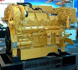 Caterpillar engine. On the right, there is a cylindrical oil filter and on slightly below the middle left also two oil filters side by side. "CAT 1R-1808 - Advanced" reads on these filters. On the bottom, it reads "CAT C32 MARINE ENGINE". On the upper right there is a black plate reading "CAT C32" along with some unreadable text. Several white quality check stickers are glued on the engine.