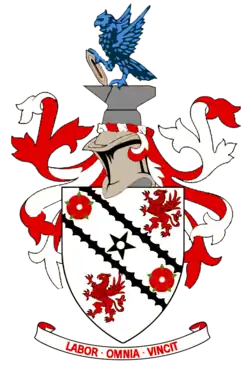 A white shield upon which emblems of two red roses and two red griffins appear at alternate corners. A black five-pointed star is in the centre of the shield's design between two black diagonal lines. Around the shield are red and white ribbons in a symmetrical design. Above the shield is a silver-coloured knight's helmet surmounted by grey anvil upon which a vivid azure-coloured eagle is perched, holding a grey shuttle. Below the shield is the motto "LABOR OMNIA VINCIT".