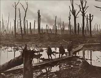 A group of soldiers walk across wooden duckboards that have been constructed over a waterlogged and muddy field. Shattered trees dot the landscape, with a low-lying haze in the background.