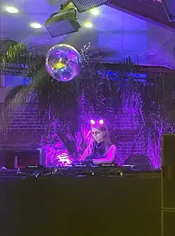 Chelsea Manning, wearing glowing cat ears, working behind a DJ booth with a discoball overhead