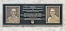 A bronze plaque that has engravings of two deceased guards at the Capitol. The text on the plaque reads: "IN HONOR AND REMEMBRANCE OF THE HEROISM DISPLAYED BY OFFICER JACOB JOSEPH CHESTNUT AND DETECTIVE JOHN MICHAEL GIBSON UNITED STATES CAPITOL POLICE WHO, ON JULY 24, 1998, HERE BRAVELY GAVE THEIR LIVES DEFENDING THE UNITED STATES CAPITOL DEDICATED BY THE HONORABLE J. DENNIS HASTERT, SPEAKER OF THE HOUSE OF REPRESENTATIVES, AND THE HONORABLE STROM THURMOND, PRESIDENT PRO TEMPORE OF THE UNITED STATES SENATE".