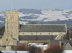 Side view of the upper section and tower of a long, flint-built church with snow-covered hills in the background and various rooftops in the foreground. A tall tower rises on the left.