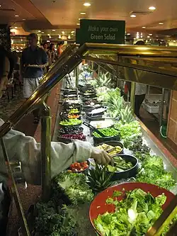 A chilled serving table with 2 rows of salad ingredients. Additional leafy and cruciferous vegetables are displayed alongside the food as decorations. A sign instructs: "Make your own Green Salad".