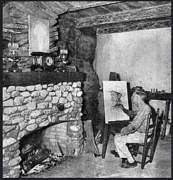 Man painting at an easel in a rustic cabin interior with exposed beams and stone fireplace