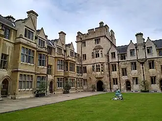 Grass lawn with small statue in centre, surrounded by neo-Gothic stone buildings with Elizabethan bay windows