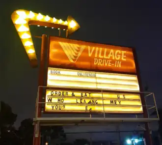 Illuminated entrance sign to Coburg Drive-In cinema. A giant arrow of lights invites cars in. The letters are jumbled up into nonsense. The sign reads "BOOK ONLINENOW. ORDER & EAT US W HO LEA ANG MR Y YOUR EA", a corruption of "Book Online Now. Order and Eat Without Leaving Your Seat."