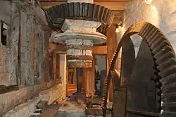 on the floor below the stones is the drive from the wheel. We see the same vertical shaft that goes up through the floor, topped here with a black iron bevel gear. Another bevel gear on the right, currently disengaged, is driven by the waterwheel. The room is cramped and whitewashed.