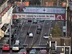 High level shot of multi lane road lower down stretching into distance with two glass sided pedestrian walkways over linking car park to right and shopping centre to left