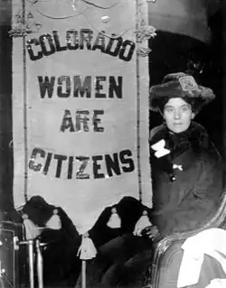 A young woman suffragette sits in a chair near a banner reading "Colorado Women are Citizens." The woman wears a fur coat, hat, and leather gloves. Tassels dangle from the top and bottom of the banner.