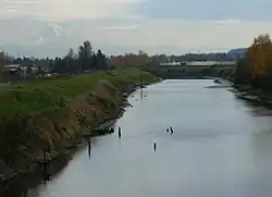 A stream perhaps 50 feet (15 m) wide flows between raised, grass-covered banks on either side. Several poles or sticks, perhaps remnants of a derelict structure, poke above the water in places. Low buildings and many trees are in the middle distance. A mountain range dominated by a snow-covered peak is visible in the far distance.