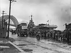 Black and white photo of a dockside scene. Buses are driving in front a crowd of people. An aircraft carrier and a bridge are visible in the background.