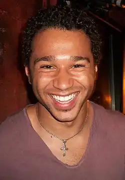 Headshot of Corbin Bleu smiling. He is a young, black man with curly hair and stubble, wearing a plain light red shirt and a cross necklace.