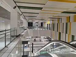 The interior of Hume station with a color scheme of green, yellow, and white. The walls and ceiling are decorated with panels in these colors. An escalator leads down from the concourse to the lower platform level.