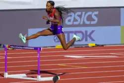 Photo of Dalilah Muhammad while jumping over a hurdles on red athetics tracks