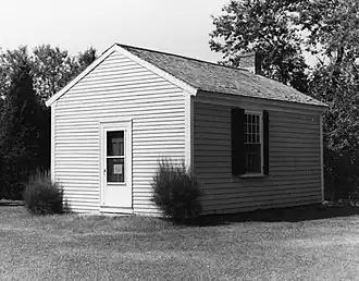 A black and white photograph of a small one-room building. It has a door on one side and a window on another.