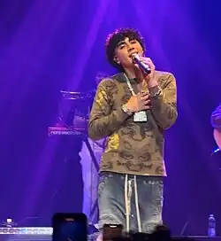 DannyLux performing at Syracuse University on October 19th, 2024. He is wearing an army-green shirt, a chain with diamond encrusted castle, blue pants, and diamond earrings