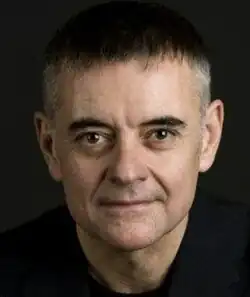 Dramatic colour publicity shot against black background, of a clean-shaven, unsmiling man with dark hair, brushed forward..