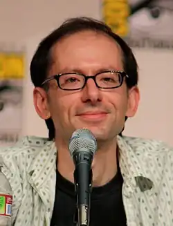 A closeup of a man in front of a microphone. He has a receding hairline and wears dark-framed glasses.