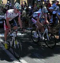 A pair of cyclists on the road, wearing matching white jerseys with blue and red trim. A car is partly visible behind them, and numerous spectators are on the roadside.