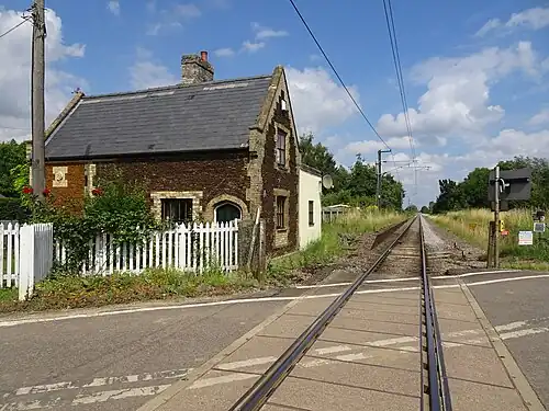 A photograph taken from a level crossing above a single railway track. To the left is a Tudor revival brick building with a slate roof, and the shape of a platform next to the railway but with no surface.