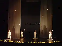 Four women in straight, white dresses stand on a black stage with a row of candles at the front edge. Diaphanous curtains hang from the ceiling between the first and second and the third and fourth women. There are hanging light bulbs and a screen behind them reads "7. Desdemona/Othello".