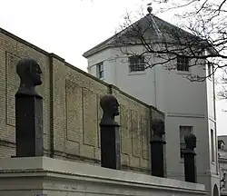 Four evenly spaced sculptures of bald male heads with slightly different facial expressions, each standing on a black rectangular plinth on a flat white surface in front of a yellow-brick wall and an octagonal grey-roofed tower.