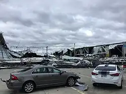 Two cars sit in the parking lot in front of the destroyed factory building, which has lost its roof in the image.