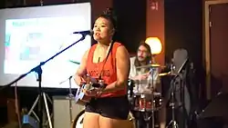 Person of Asian descent wearing a orange tank top and standing on stage singing into a microphone and strumming a wooden guitar