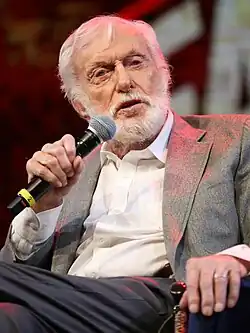 A white-haired man wearing a grey suit and white shirt talks with a microphone