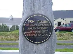 Black circular plaque mounted on a standing rock. Text reads: "Don Allum, the first man to row across the Atlantic in both directions, landed on the rocky shore down from this memorial, from St. John's Newfoundland on 4th September 1987, completing the second leg of his epic voyage in a 19 ft. 10 ins. plywood open boat, the QE 3, after 77 days at sea."