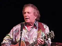 A grey-haired man in a brightly patterned shirt playing a guitar and singing into a microphone