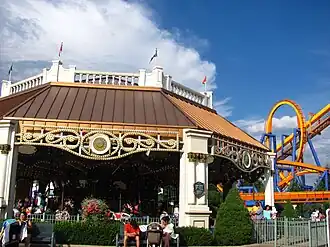 A shelter building with a bronze roof containing a historic carousel with a yellow and blue roller coaster in the background