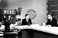 Two men sitting behind a desk next to a camera with "WCKT" and "channel 7" signage on it. Behind one of the men is a logo for Miami Dade Junior College.