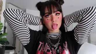 Headshot of a female YouTuber as an e-girl posing with her tongue out. She is a young white woman with long, dark hair in buns, wearing heavy make-up and street fashion clothing.