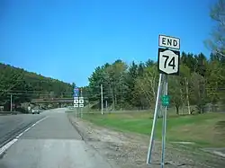 An intersection of a pair of highways in a wooded rural area. In the foreground is a sign assembly reading "End NY 74"; in the foreground is a second assembly indicating that Interstate 87 is straight ahead and US 9 is accessed by turning either left or right.